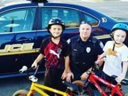 2 children pose with a police officer in front of a police car with their bicycles