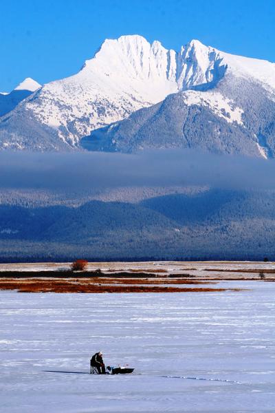 Ice fisherman sitting on a frozen flathead lake with snow covered mountain in the background 