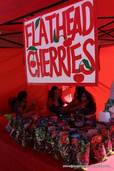 Children sitting in a red stand with bags of cherries for sale with a sign above that reads "Flathead Cherries"