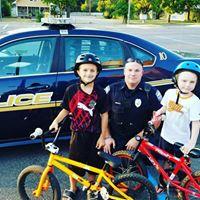 2 children pose with a police officer in front of a police car with their bicycles