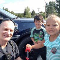 Police officer posing with two children on their scooters in front of a police car